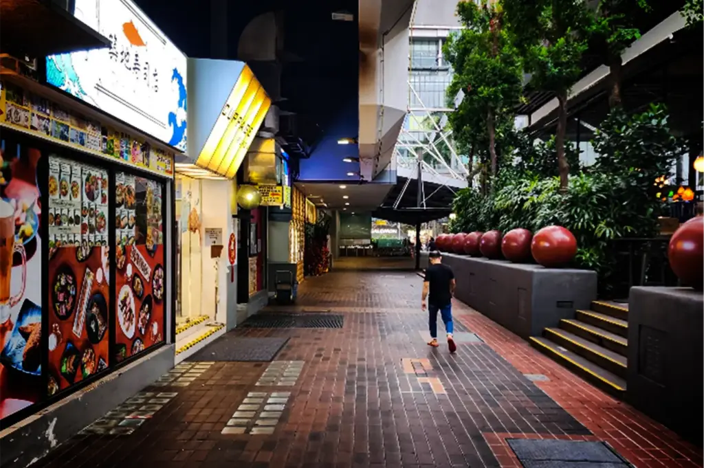 An eye-level, medium-wide shot captures a quiet pedestrian walkway stretching alongside a commercial building at night, representing the file "4-Cuppage Plaza Singapore A Quiet Food Landmark.webp". On the left, illuminated storefronts feature large, colorful food menus plastered on glass windows, topped by glowing signs including one with a yellow awning for Cuppage Plaza. A lone pedestrian in a black t-shirt and blue jeans walks down the center of the brick-paved corridor away from the camera. The right side of the path is bordered by a low grey concrete wall topped with a row of large, decorative red spherical ornaments, which separates the walkway from an elevated patio area filled with lush green trees and potted plants, creating a calm and modern urban atmosphere.