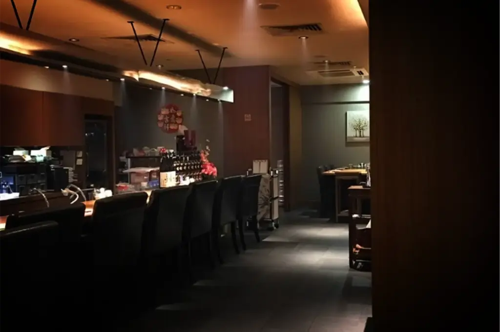 An eye-level, medium shot captures the quiet, dimly lit interior of an elegant Japanese restaurant or sushi bar, representing the file "5-Beyond the Usual 5 Hidden Cuppage Plaza Food.webp". On the left, a long, curved dining counter is lined with a row of high-backed black leather chairs, facing a service area stocked with bottles of sake, small dishes, and a floral arrangement. Spotlights embedded in the warm-toned ceiling illuminate the counter area, while a dark tiled floor leads toward a separate dining table in the background, where a painting of trees hangs on a grey wall. The foreground on the right features a cropped wooden architectural column, adding a sense of depth and privacy to the sophisticated, intimate dining space.