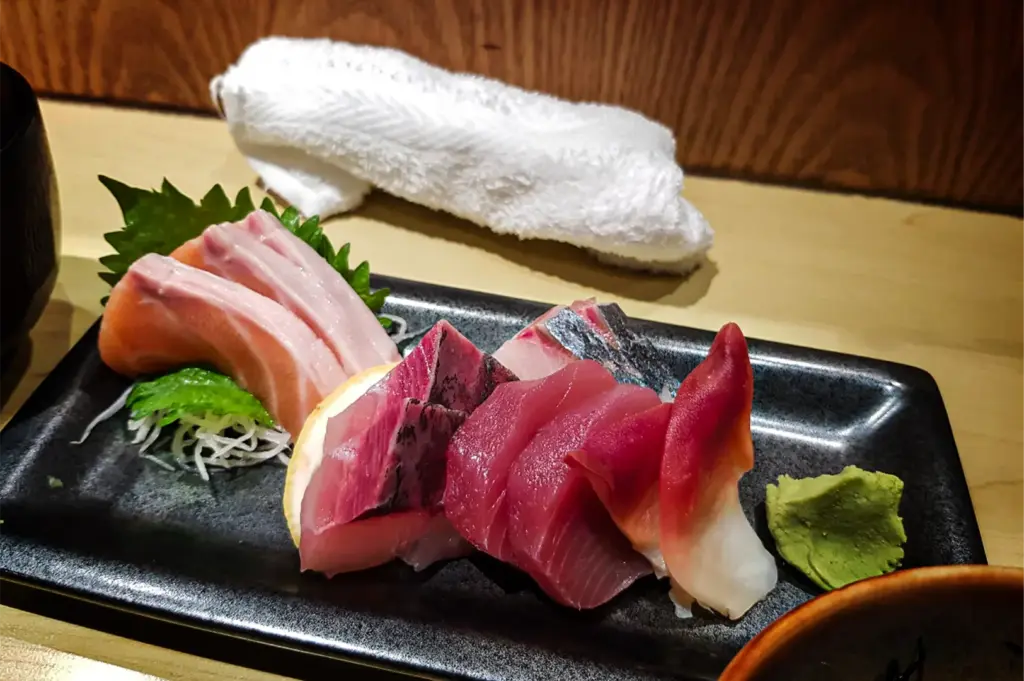 A close-up, eye-level shot captures a beautifully arranged platter of fresh assortment sashimi on a long, rectangular black plate, representing the file "5-Cuppage Plaza Singapore A Quiet Food Landmark.webp". On the left, thick slices of pale pink and orange salmon belly sit elevated on green shiso leaves and a bed of shredded daikon radish. Moving right, the platter features rich, reddish pink slices of tuna (maguro), silver-skinned mackerel (saba), and a piece of red-tipped surf clam (hokkigai). A small dollop of bright green wasabi paste rests on the far right of the plate, while a rolled white hand towel (oshibori) sits on the light wooden counter background against a warm wooden wall panel, altogether evoking a premium and authentic Japanese dining experience.