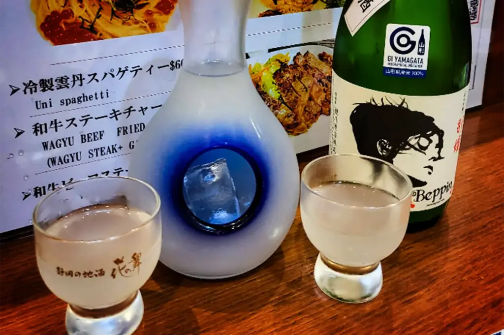 A close-up, eye-level shot captures a traditional Japanese sake serving set arranged on a wooden table, representing the file "5-Keria Japanese Restaurant.webp". In the center stands a frosted glass sake carafe (tokkuri) filled with a slightly cloudy, milky-white sake; the carafe features a distinctive, deep-blue hollow center chamber holding ice cubes to keep the beverage chilled. Flanking the carafe are two small, clear glass sake cups (ochoko) filled with the same cloudy sake, with the left cup adorned with delicate Japanese calligraphy. To the right sits a green glass sake bottle featuring a white label with a black artistic sketch of a face and the text "GI YAMAGATA" and "Beppin," while printed menus displaying items like "Uni spaghetti" and "WAGYU BEEF FRIED" rest in the background, completing the authentic dining setting.