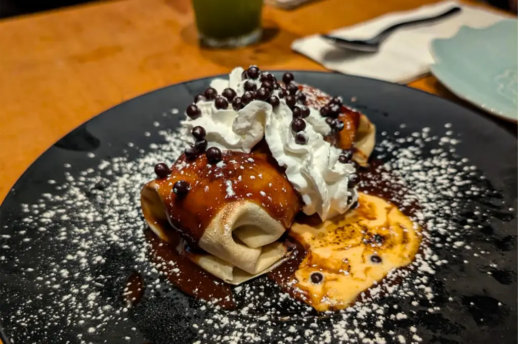 Folded Japanese-style dessert crepes drizzled with chocolate sauce and topped with whipped cream, chocolate pearls, and powdered sugar, served on a black plate in a cozy restaurant.