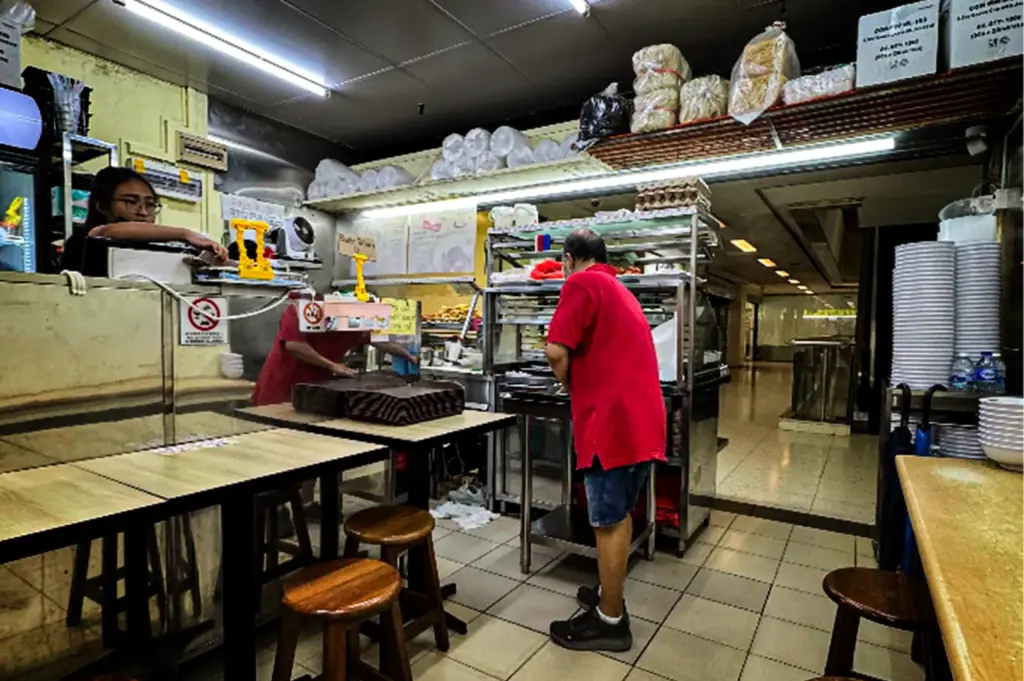 An eye-level, medium shot captures the bustling kitchen and ordering counter of a casual eatery, representing the file "6-Beyond the Usual 5 Hidden Cuppage Plaza Food.webp". In the center, a man in a bright red polo shirt and denim shorts stands with his back partially to the camera, working near a stainless steel food prep station, while another worker in a red shirt leans over a counter behind a stack of trays on the left. A staff member with glasses looks on from a raised service window in the upper left corner. The small dining space features simple wooden tables, round wooden stools, and tiled flooring, with high shelves packed with plastic bowls, wrapped ingredients, and storage boxes, looking out into a brightly lit shopping corridor in the background.