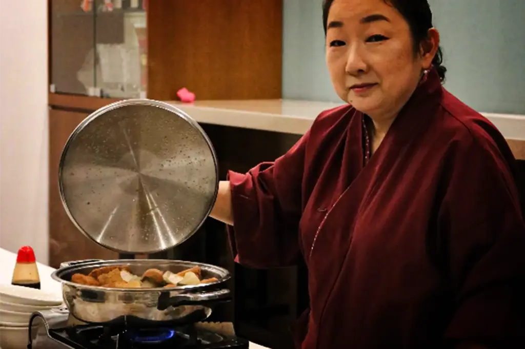 An eye-level, medium close-up shot captures a woman in a traditional dark red or maroon kimono-style wrap jacket presenting a hot dish, representing the file "6-Keria Japanese Restaurant.webp". Looking directly toward the camera with a subtle, pleasant expression, she holds up a large, circular stainless steel lid covered in condensation to reveal a steaming hot pot meal cooking inside a shallow metal pot over a small blue flame stove. The simmering dish contains visible chunks of tofu, fish cakes, or root vegetables, while a small stack of white bowls and a soy sauce dispenser rest on the white table surface to the far left against a soft, out-of-focus background.