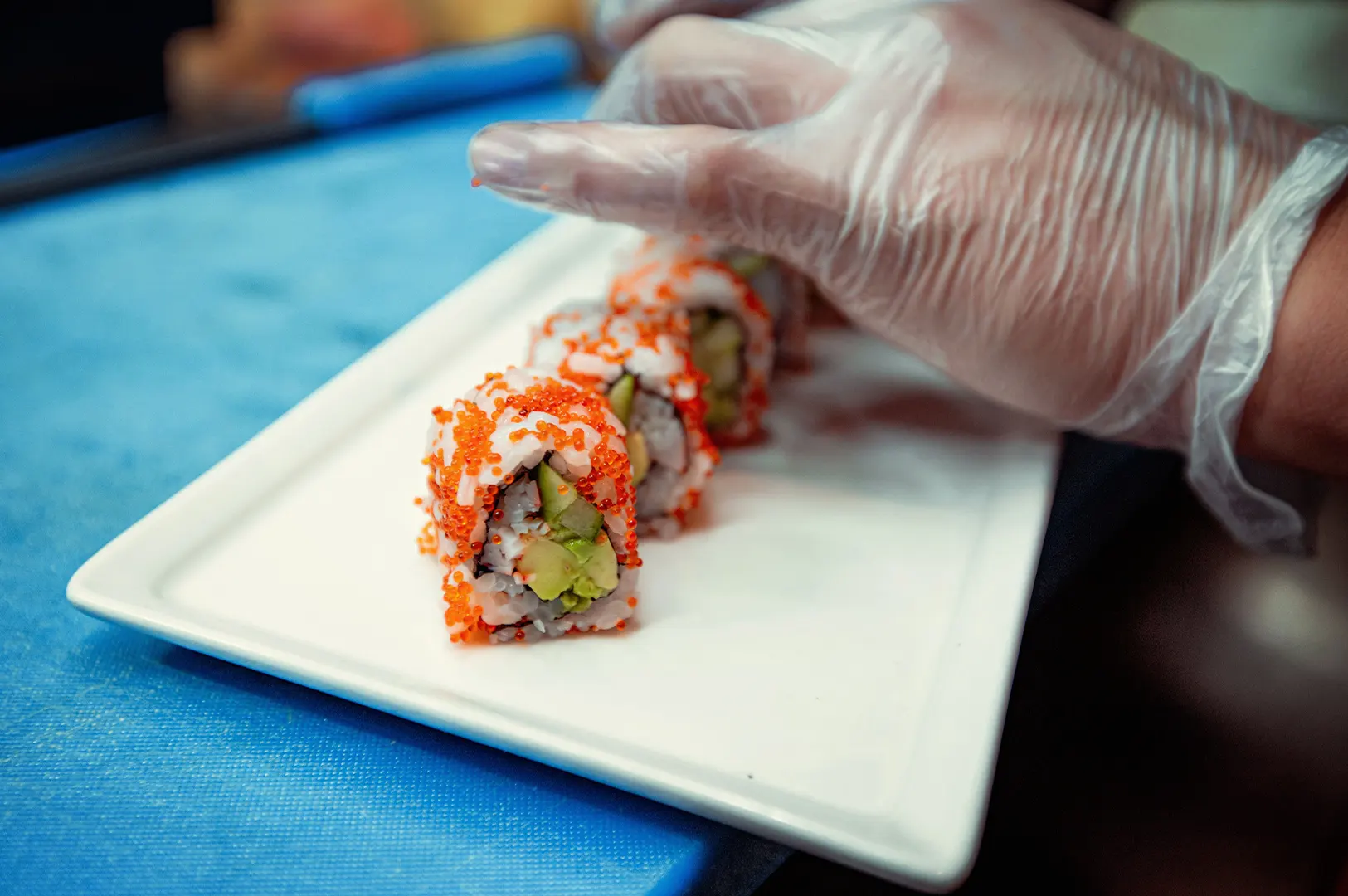 A close-up shot captures a chef wearing clear plastic gloves carefully arranging a row of sushi rolls on a long, rectangular white plate. The inside-out rolls are filled with avocado and cucumber, coated in vibrant orange tobiko, and set against a bright blue countertop.