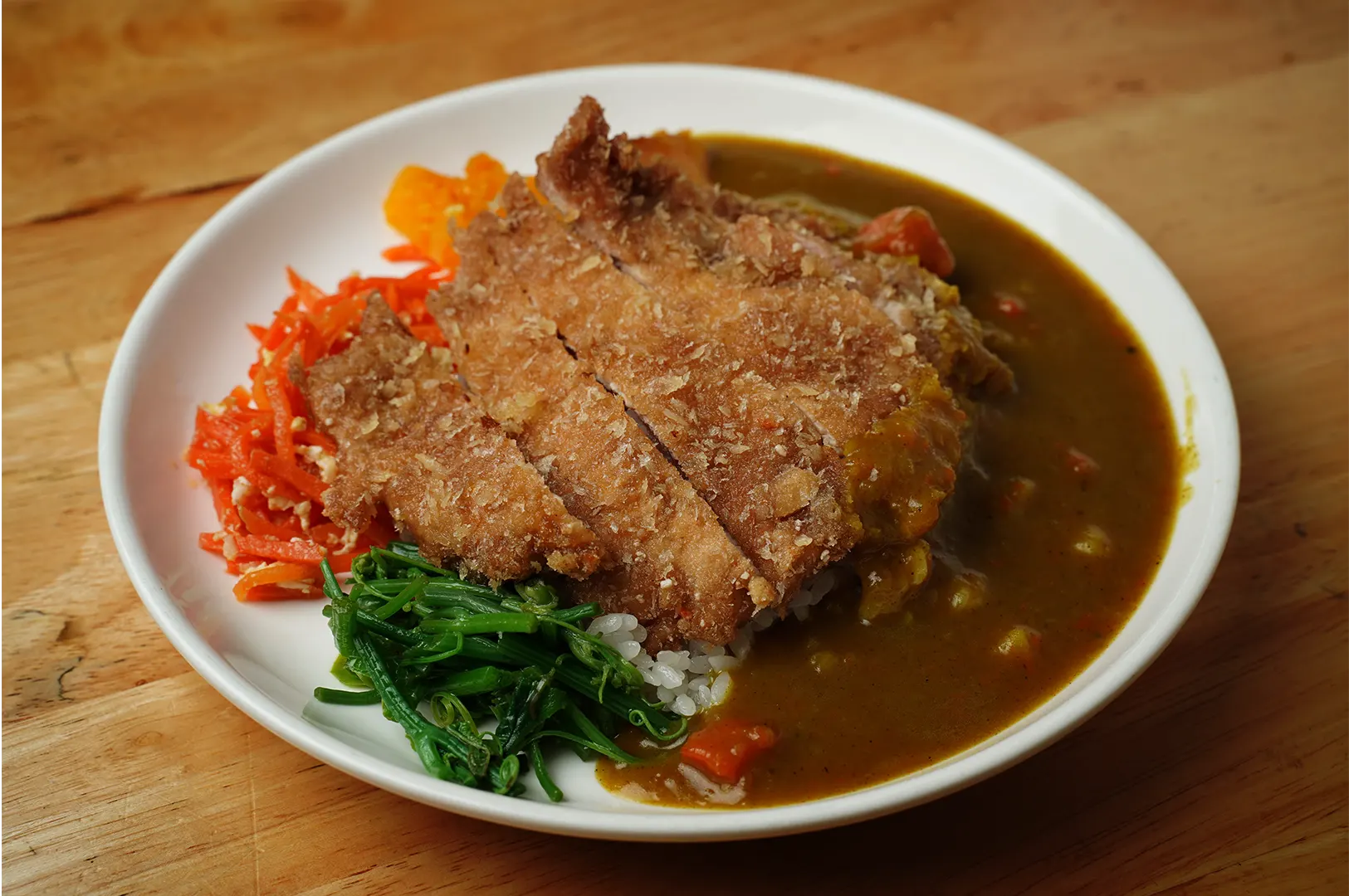 A high-angle, close-up shot captures a vibrant plate of Japanese chicken or pork katsu curry, representing the file "1-A First-Timer’s Guide to Japanese Curry in Singapore.webp". Served in a shallow white ceramic bowl on a light-colored wooden table, a golden-brown, crispy deep-fried cutlet is sliced and angled across a bed of white rice. The right side of the plate is filled with a rich, glossy brown curry sauce containing small chunks of carrots, while the left side features a colorful assortment of side dishes, including a mound of bright orange shredded carrots, a small bundle of cooked green leafy vegetables, and a spoonful of yellowish pickled radish, creating a wholesome and appealing meal presentation.