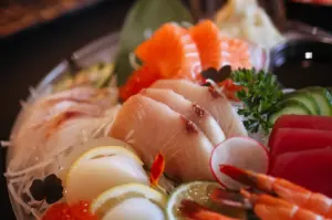 A vibrant, close-up shot captures an assortment of fresh sashimi beautifully arranged on a bed of shredded daikon radish. The platter features thick slices of salmon, tuna, and white fish alongside scallops with lemon garnishes, sweet shrimp, cucumber slices, and a small mound of salmon roe.