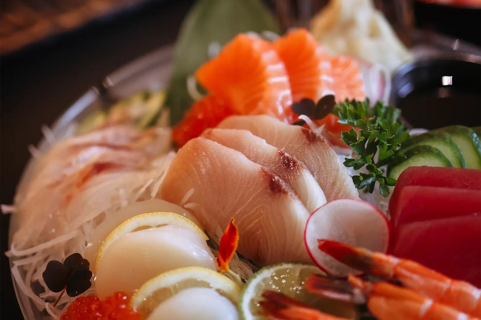 A vibrant, close-up shot captures an assortment of fresh sashimi beautifully arranged on a bed of shredded daikon radish. The platter features thick slices of salmon, tuna, and white fish alongside scallops with lemon garnishes, sweet shrimp, cucumber slices, and a small mound of salmon roe.