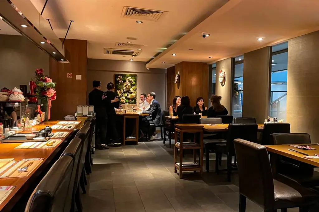 A wide, eye-level shot captures the warm and intimate dining room of a modern Japanese restaurant, representing the file "1-Beyond the Door Savoring Tranquility at Hanashizuku.webp". On the left, an empty wooden bar counter lined with dark leather high-backed chairs sits neatly set for service with small bowls and placemats, decorated with a vibrant floral arrangement at the far end. In the center and background, several small groups of patrons are seated at wooden dining tables engaged in conversation, while attentive staff members in dark uniforms stand nearby to assist them. The space features grey tiled flooring, wood-paneled accents, a textured moss-and-floral art piece on the back wall, and soft, recessed ceiling lighting that creates a relaxed and upscale ambiance.