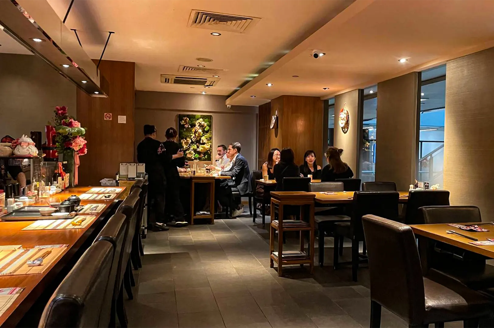 A wide, eye-level shot captures the warm and intimate dining room of a modern Japanese restaurant, representing the file "1-Beyond the Door Savoring Tranquility at Hanashizuku.webp". On the left, an empty wooden bar counter lined with dark leather high-backed chairs sits neatly set for service with small bowls and placemats, decorated with a vibrant floral arrangement at the far end. In the center and background, several small groups of patrons are seated at wooden dining tables engaged in conversation, while attentive staff members in dark uniforms stand nearby to assist them. The space features grey tiled flooring, wood-paneled accents, a textured moss-and-floral art piece on the back wall, and soft, recessed ceiling lighting that creates a relaxed and upscale ambiance.