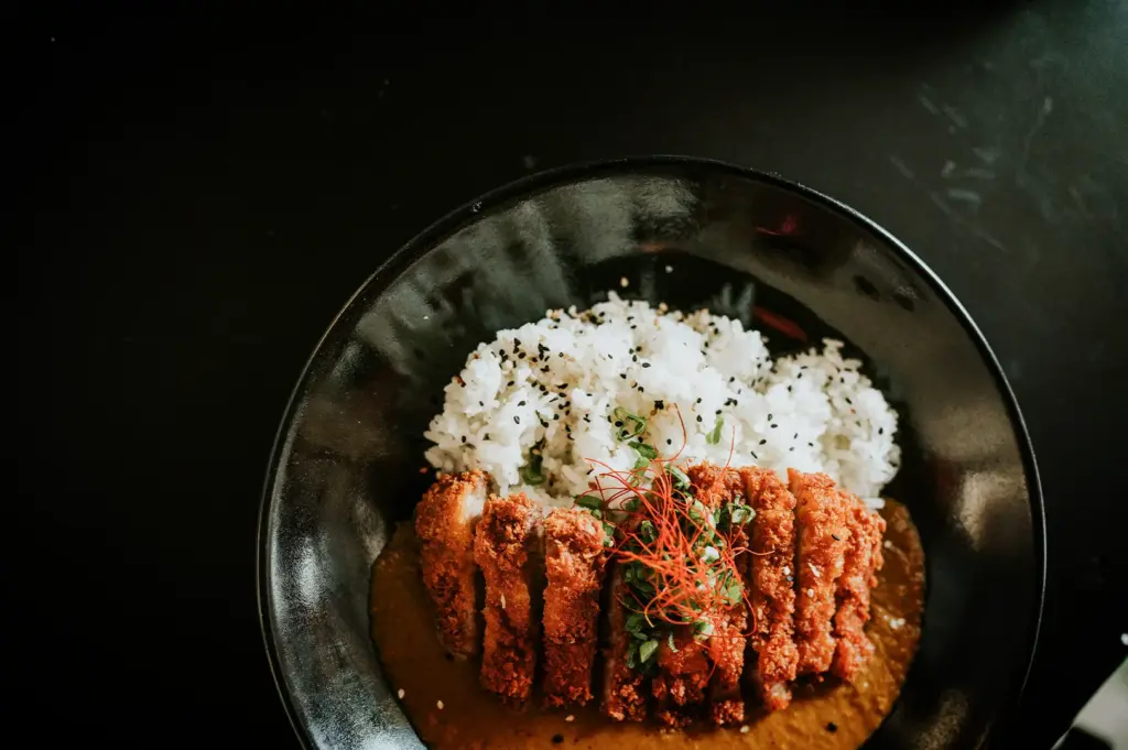 An overhead, top-down shot showcases a beautifully presented Japanese katsu curry dish served in a large black ceramic bowl, representing the file "1-From Hidden Counters to Iconic Eateries 5 Best Japanese Curry.webp". The bowl is cleanly divided into two sections: the top half holds a mound of fluffy white rice sprinkled with black sesame seeds, while the bottom half contains a rich, thick, golden-brown curry sauce. Resting perfectly across the center is a sliced, crispy chicken or pork katsu cutlet with a deep golden-brown panko crust. The cutlet is artfully garnished in the center with a scatter of finely chopped green scallions and a delicate cluster of thin red chili threads, all set against a stark, minimalist black background that emphasizes the vibrant colors of the meal.
