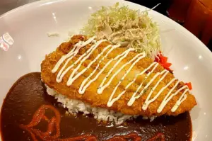 A high-angle, close-up shot captures a massive serving of Japanese katsu curry in a colossal white ceramic dish, representing the file "1-Monster Curry and the Meal That Fills the Table.webp". A giant, golden-brown fried fish or chicken cutlet with a wonderfully crispy breading lies across a bed of white rice, elegantly drizzled with zig-zagging lines of white mayonnaise and a tiny pinch of parsley. The cutlet sits in a vast pool of thick, dark brown curry sauce, which features a decorative swirl of reddish chili sauce in the lower-left corner. A mound of finely shredded cabbage tossed in a creamy sesame dressing and a small heap of red pickled radish (fukujinzuke) are tucked into the upper right background, completing the signature, oversized meal presentation.