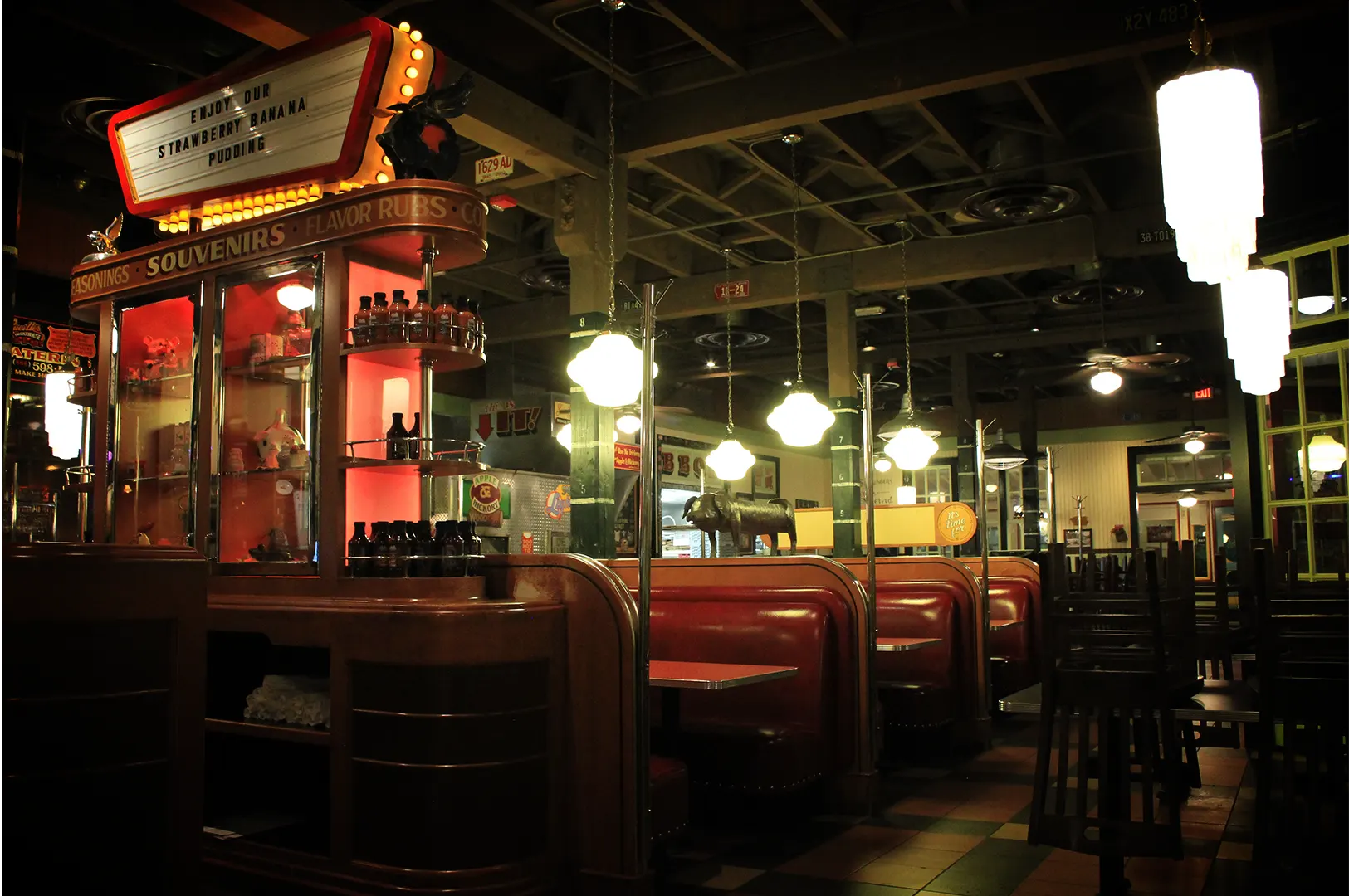 A wide-angle, eye-level indoor shot captures the nostalgic, vintage interior of an American diner or barbecue restaurant, representing the file "1-Past the Last Familiar Turn.webp". In the foreground on the left, a large wooden display cabinet stands out with glowing red accent panels, displaying rows of sauce bottles and "SOUVENIRS" labeling. Atop the cabinet sits an illuminated retro marquee sign that reads, "ENJOY OUR STRAWBERRY BANANA PUDDING." Extending into the background, a series of classic red leather booths are arranged under a row of hanging milk-glass pendant lights, while a small metal pig statue rests on a partition wall. The restaurant features an open ceiling structure with industrial beams, ceiling fans, and stacked frosted glass light fixtures on the right, creating a warm, moody, and dimly lit dining atmosphere.