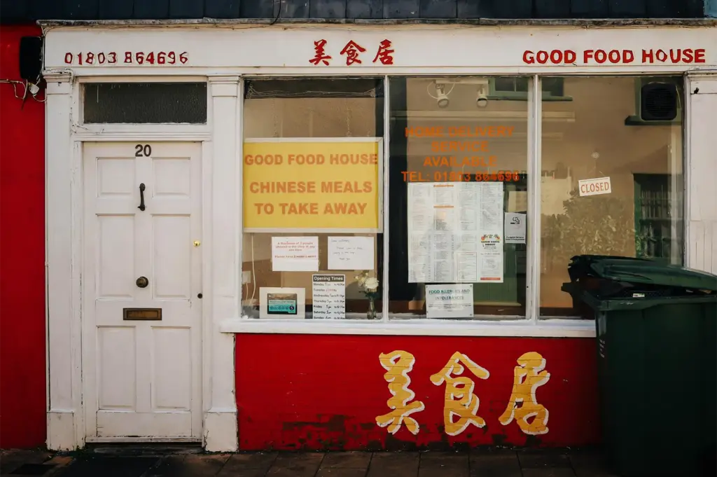 A straight-on, eye-level shot captures the charming and weathered storefront of a Chinese takeaway restaurant, representing the file "1-The Longer Way Home.webp". On the left, a white residential-style front door marked with the number "20" sits next to a large display window framed in white trim. The shop features bright red accents on the lower brickwork and left wall, adorned with large yellow Chinese characters, which are mirrored in red script on the white signboards above alongside the English name "GOOD FOOD HOUSE" and a phone number. The large glass window reveals a prominent yellow sign reading "CHINESE MEALS TO TAKE AWAY," an extensive printed menu, a small "CLOSED" sign dangling in the corner, and a glimpse of a dark green trash bin parked on the sidewalk to the far right, evoking a quiet, nostalgic street corner scene.