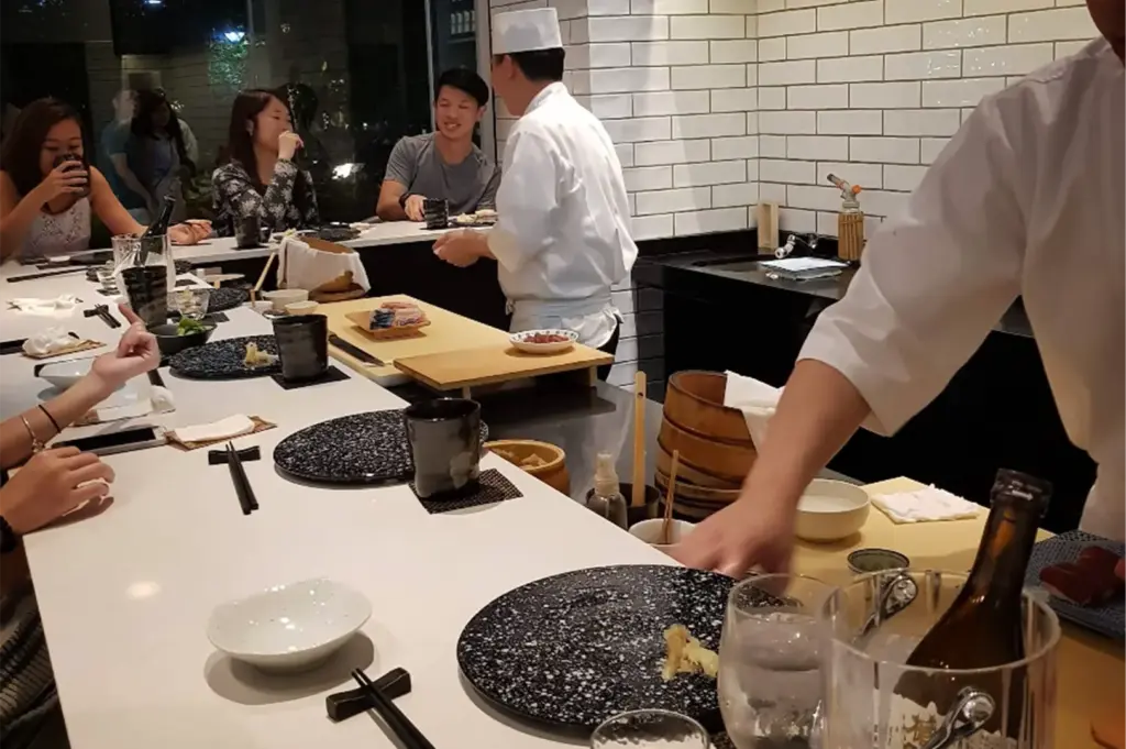 An eye-level shot captures a lively omakase sushi bar where chefs clad in traditional white uniforms prepare fresh dishes behind a sleek white counter. Several patrons sit along the bar chatting and enjoying drinks, surrounded by an array of ceramic tableware, wooden preparation boards, and stacked bamboo rice barrels.