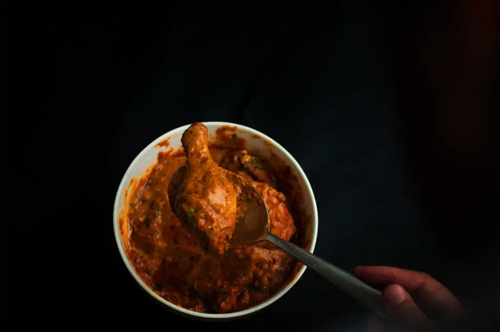 A top-down, dramatic close-up shot captures a portion of rich curry being served, representing the file "2-A First-Timer’s Guide to Japanese Curry in Singapore.webp". Set against a stark black background, a white ceramic bowl is filled with a thick, textured, reddish-orange curry sauce flecked with herbs. In the center, a hand uses a large metal spoon to lift a succulent piece of bone-in chicken—resembling a drumstick—heavily coated in the thick gravy, creating a high-contrast and mouth-watering focal point.