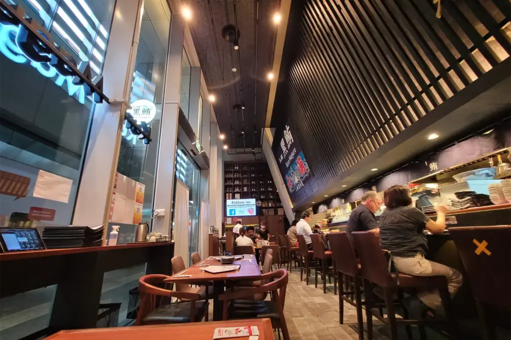 A low-angle shot captures the modern, high-ceiling interior of a Japanese restaurant, featuring a dining bar on the right lined with patrons seated beneath a prominent wooden-slat wall accent. To the left, floor-to-ceiling glass windows overlook the street next to a row of empty wooden tables and chairs.