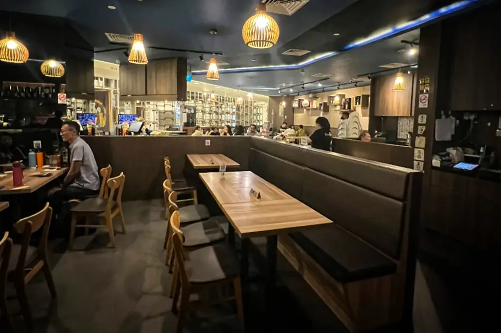 An eye-level shot captures the warmly lit interior of a contemporary restaurant featuring long wooden dining tables paired with cushioned booth seating and wooden chairs. The ambient space is illuminated by woven globe pendant lights hanging from a dark ceiling, with patrons dining and staff working in the background.