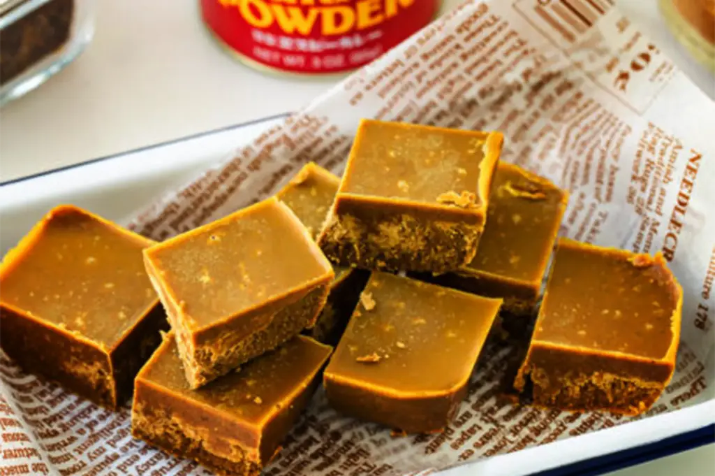 A close-up, slightly high-angle shot captures a small pile of golden-brown Japanese curry roux blocks, representing the file "3-A First-Timer’s Guide to Japanese Curry in Singapore.webp". The thick, dense spice cubes are stacked neatly inside a shallow white container lined with newspaper-printed parchment paper, showcasing their smooth top surfaces and slightly textured, crumbly undersides. In the softly blurred background, the red lid of a curry powder tin is partially visible, hinting at the culinary ingredients used to make a rich curry base.