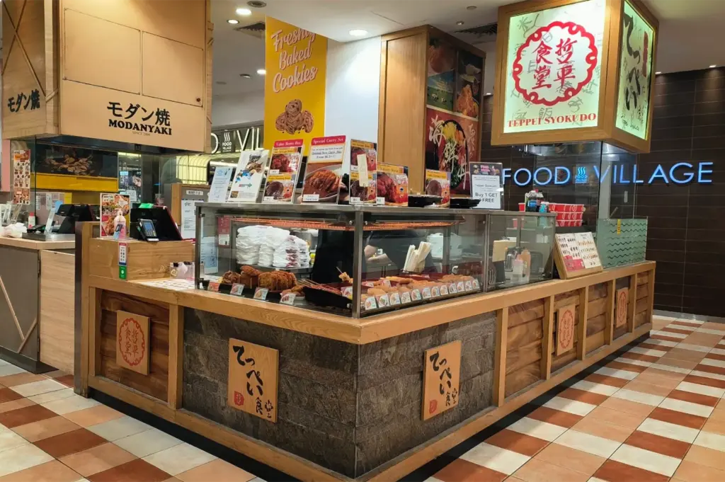 An eye-level shot captures a Japanese food stall named "Teppei Syokudo" located within a brightly lit indoor food village mall corridor. The stall features a wooden and stone counter with glass display cases showing prepared fried foods, topped with multiple colorful menu signs and a large illuminated box sign overhead.