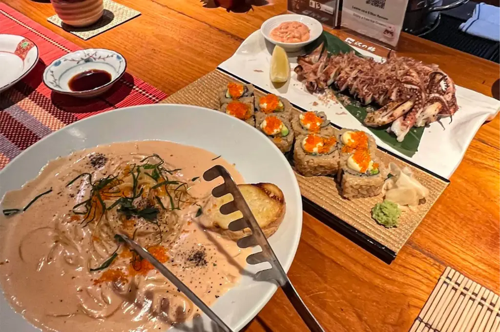 A high-angle, close-up shot captures a variety of Japanese-inspired dishes spread across a warm wooden dining table. In the foreground, a large white bowl holds creamy mentaiko pasta topped with orange roe and shredded nori, accompanied by a piece of toasted bread and a metal serving tong. Behind it, a long wooden board presents several pieces of maki sushi topped with dollops of mayonnaise and bright orange roe, served with a side of wasabi and pickled ginger. To the right, a large white platter features a grilled whole squid sliced into sections and topped with dancing bonito flakes, served with a lemon wedge and a small dish of creamy dipping sauce, all set against a background of red woven placemats and small condiment dishes.