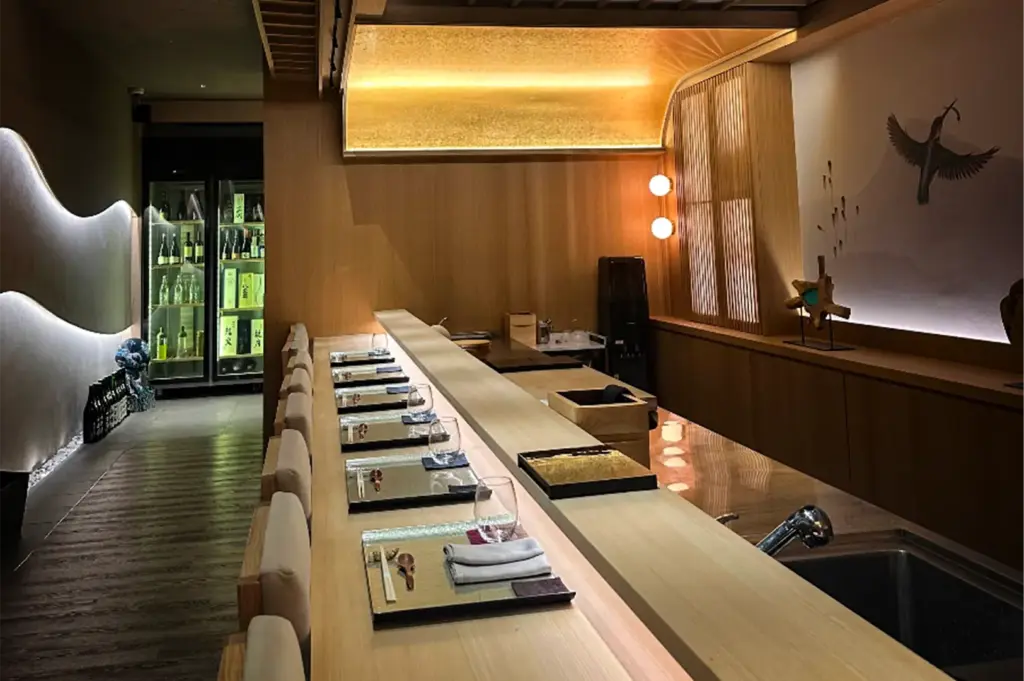An eye-level shot captures an empty, upscale sushi bar featuring a long wooden counter meticulously set with formal place settings, glasses, and grey napkins. The sophisticated space is illuminated by warm, golden accent lighting along a curved wooden canopy and features a wall mural of a flying bird, while a glass-door beverage cooler is visible in the background hallway.