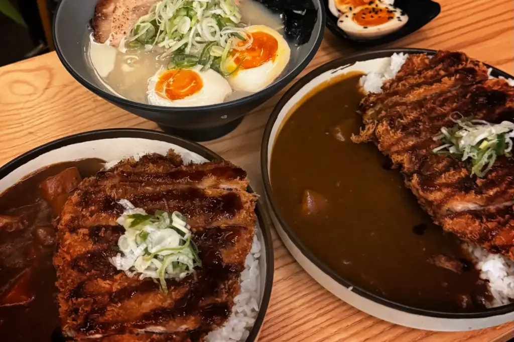 A high-angle, medium close-up shot captures a hearty spread of Japanese comfort food on a light wooden table, representing the file "4-A First-Timer’s Guide to Japanese Curry in Singapore.webp". In the foreground, two large plates of tonkatsu curry feature thick, crispy, deep-fried pork cutlets sliced and drizzled with a dark savory sauce, garnished with shredded green onions, and served alongside white rice and a rich, dark brown curry gravy. Positioned just behind them is a deep bowl of hot ramen filled with a milky broth, noodles, a slice of chashu pork, a generous mound of chopped scallions, and two soft-boiled egg halves with vibrant orange yolks, with an extra dish of seasoned ramen eggs sitting in the upper right background.