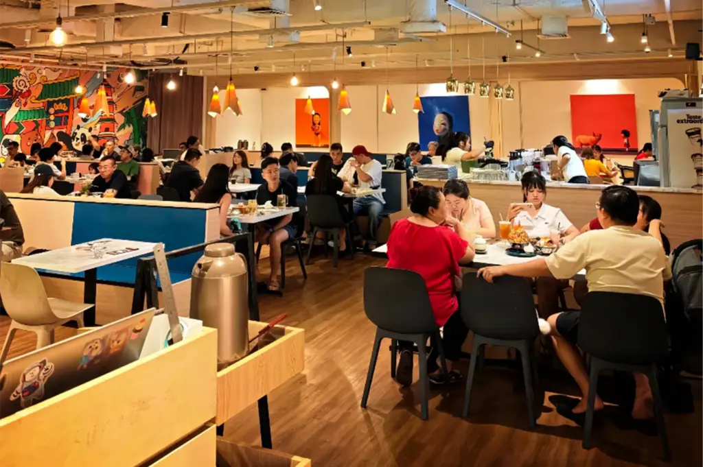An eye-level shot captures a busy, brightly lit restaurant filled with patrons dining at various wooden tables and blue booth seating. The spacious interior features warm hardwood floors, hanging pendant lights, and walls adorned with colorful, contemporary artwork and a large cartoon mural.
