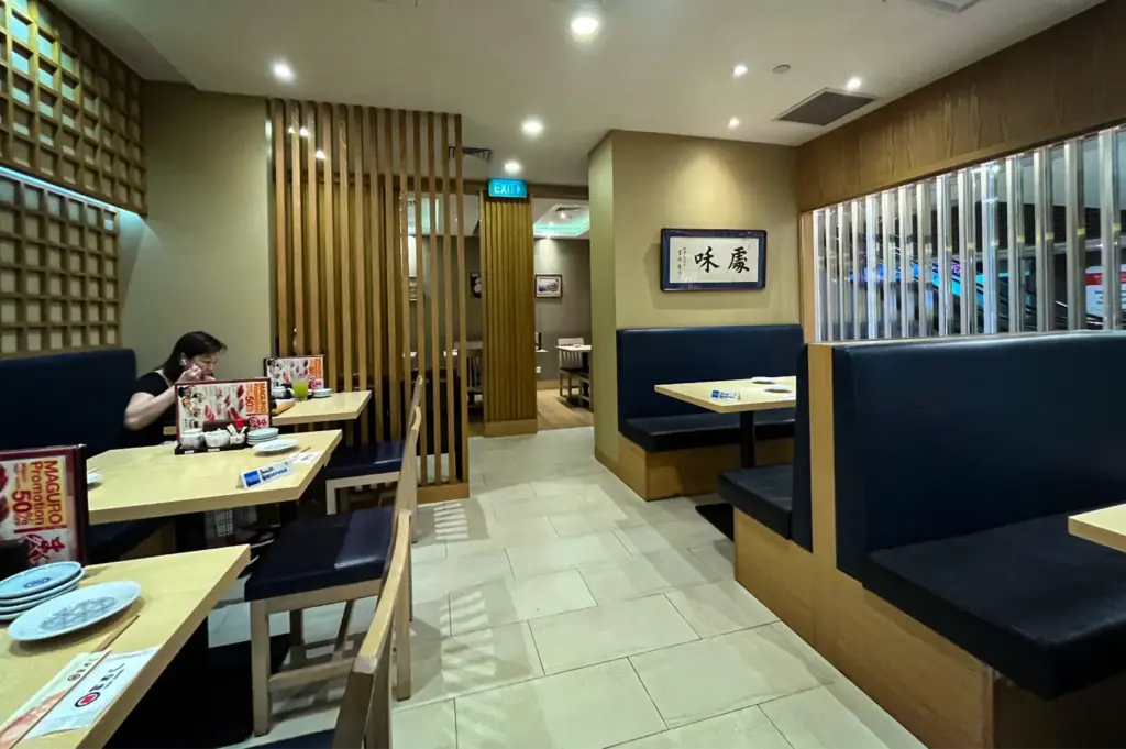 An eye-level shot captures the interior of a Japanese restaurant featuring light wood dining tables paired with dark blue cushioned booths and chairs. The neutral-toned space is accented by vertical wooden slats, a framed Japanese calligraphy sign on the wall, and glass paneling looking out toward an escalator.