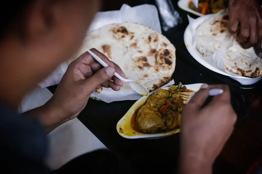 An over-the-shoulder, close-up shot captures a person eating a meal of curry and flatbread, representing the file "5-A First-Timer’s Guide to Japanese Curry in Singapore.webp". In the foreground, the diner's hands hold a white plastic spoon and fork, preparing to dig into a small, oval ceramic dish filled with a thick, savory chicken and vegetable curry. Just behind the curry dish sits a large, circular piece of charred naan or roti flatbread resting on a paper-lined plate. A shallow depth of field keeps the diner's hands and the curry bowl in sharp focus, while the diner's head on the left and another person tearing flatbread in the upper right background are softly blurred.