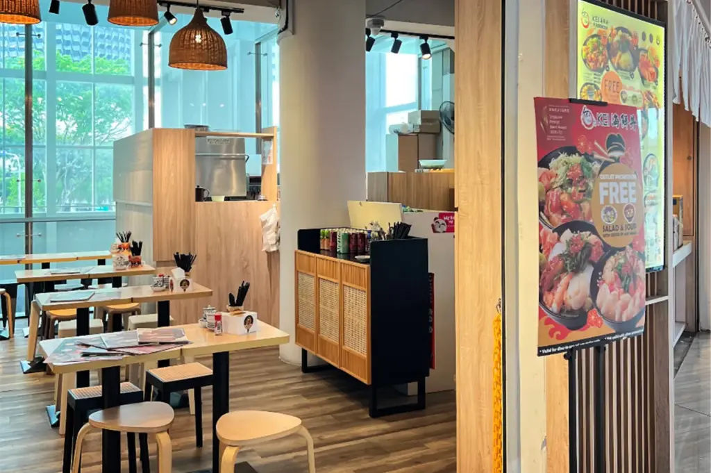 An eye-level shot captures the bright entry of a contemporary Japanese eatery, highlighted by a large promotional stand displaying colorful rice bowls next to a slatted wooden door frame. Inside, the sunlit dining area features light wood tables, minimalist stools, a service station cabinet, and woven pendant lamps hung beneath large windows.