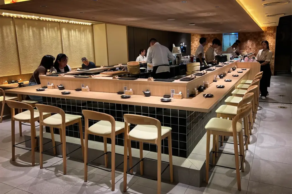 An eye-level shot captures a modern U-shaped sushi bar where chefs and staff prepare dishes for patrons seated along a light-colored wooden counter. The bar features a black grid-tiled base surrounded by matching wooden barstools with cream cushions under warm, soft interior lighting.