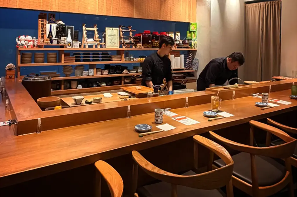An eye-level shot captures two chefs in black uniforms preparing food behind a long, polished wooden sushi counter lined with curved wooden armchairs and minimalist place settings. In the background, open wooden shelves against a deep blue wall display an assortment of ceramic bowls, plates, traditional bottles, and lucky cat figurines.
