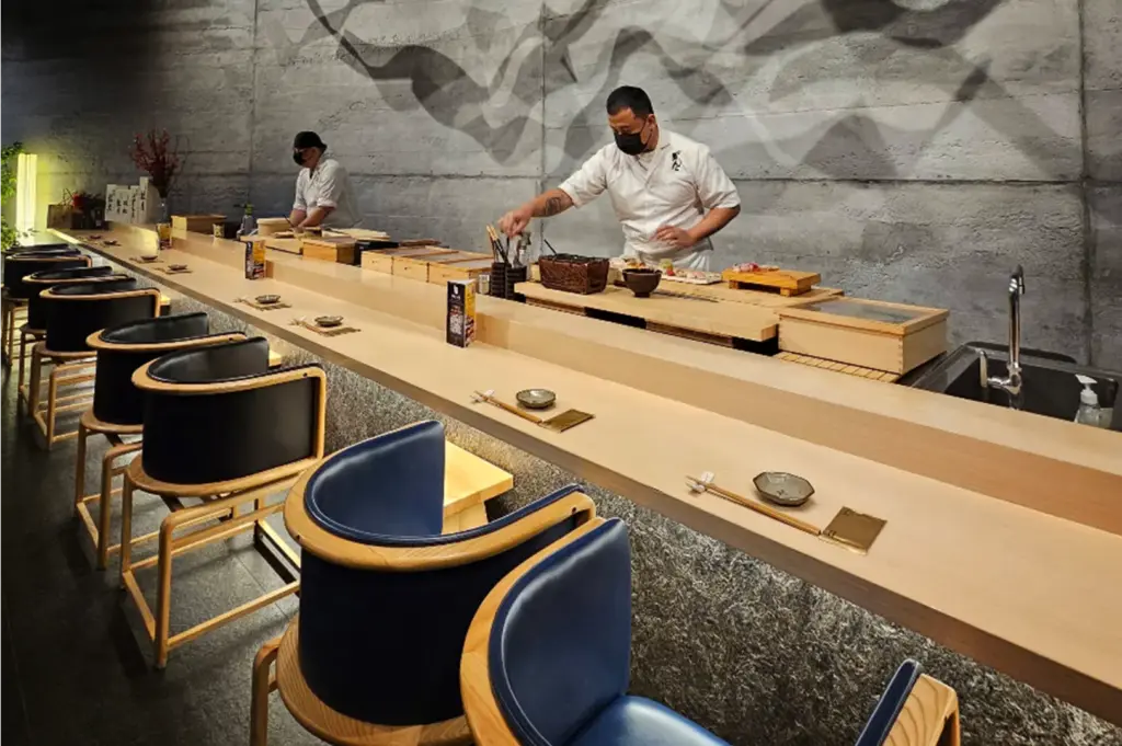 An eye-level shot captures a modern sushi bar featuring a long, light wooden counter lined with curved blue upholstered chairs and minimal place settings. Behind the counter, two chefs wearing black face masks prepare dishes at their respective wooden workstations against a textured gray concrete wall with subtle wave-like patterns.