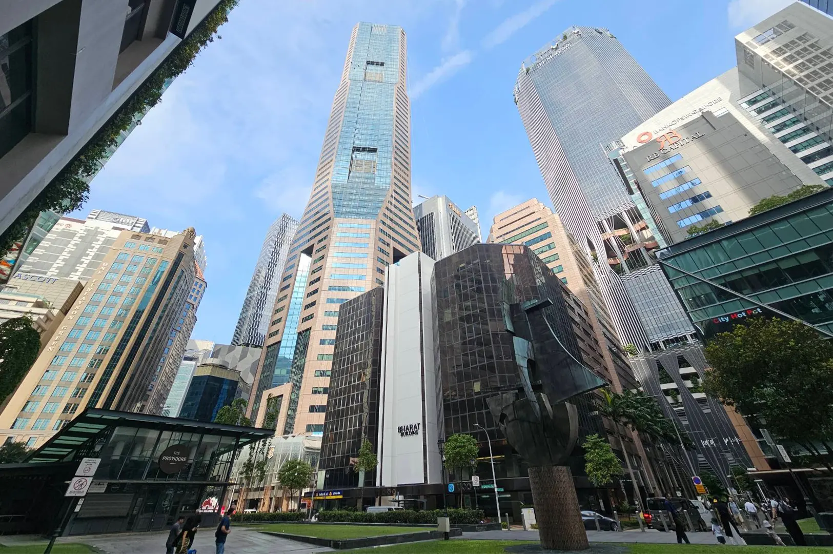 A low-angle, wide shot reveals a cluster of modern glass and stone skyscrapers, including Republic Plaza, surrounding a urban plaza under a clear blue sky. A dark metal abstract sculpture stands on the street level near green lawn patches and pedestrian walkways in the financial district.