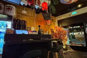 A low-angle shot captures the cozy interior of a Japanese izakaya bar, featuring a dark counter with stools, golden lucky cat figurines, and a sake fridge. The wood-paneled space is decorated with hanging paper lanterns, a traditional sake barrel, and a vibrant orange maple tree next to a large glass window.