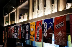 An eye-level, angled shot features a colorful string of Japanese banners adorned with kanji and festive illustrations hanging below a wooden bar structure. The shelf above is warmly lit from underneath, highlighting a collection of sake bottles neatly lined up in the background.