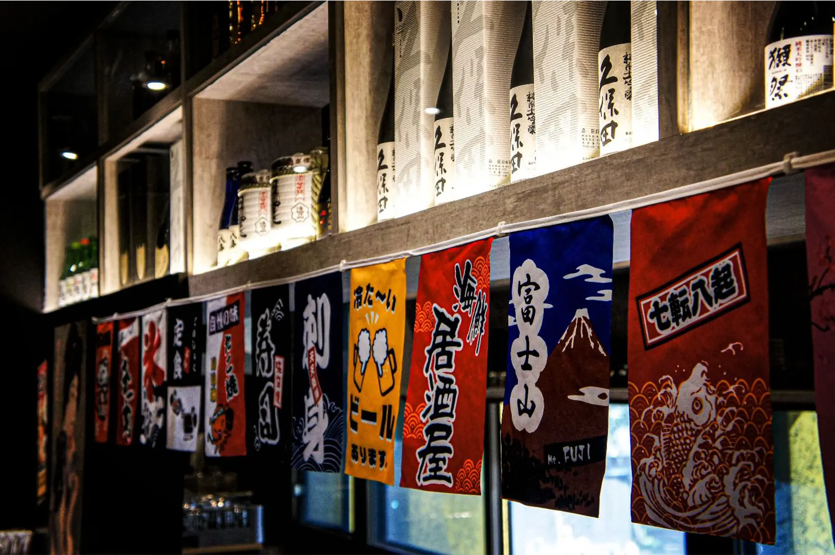 An eye-level, angled shot features a colorful string of Japanese banners adorned with kanji and festive illustrations hanging below a wooden bar structure. The shelf above is warmly lit from underneath, highlighting a collection of sake bottles neatly lined up in the background.