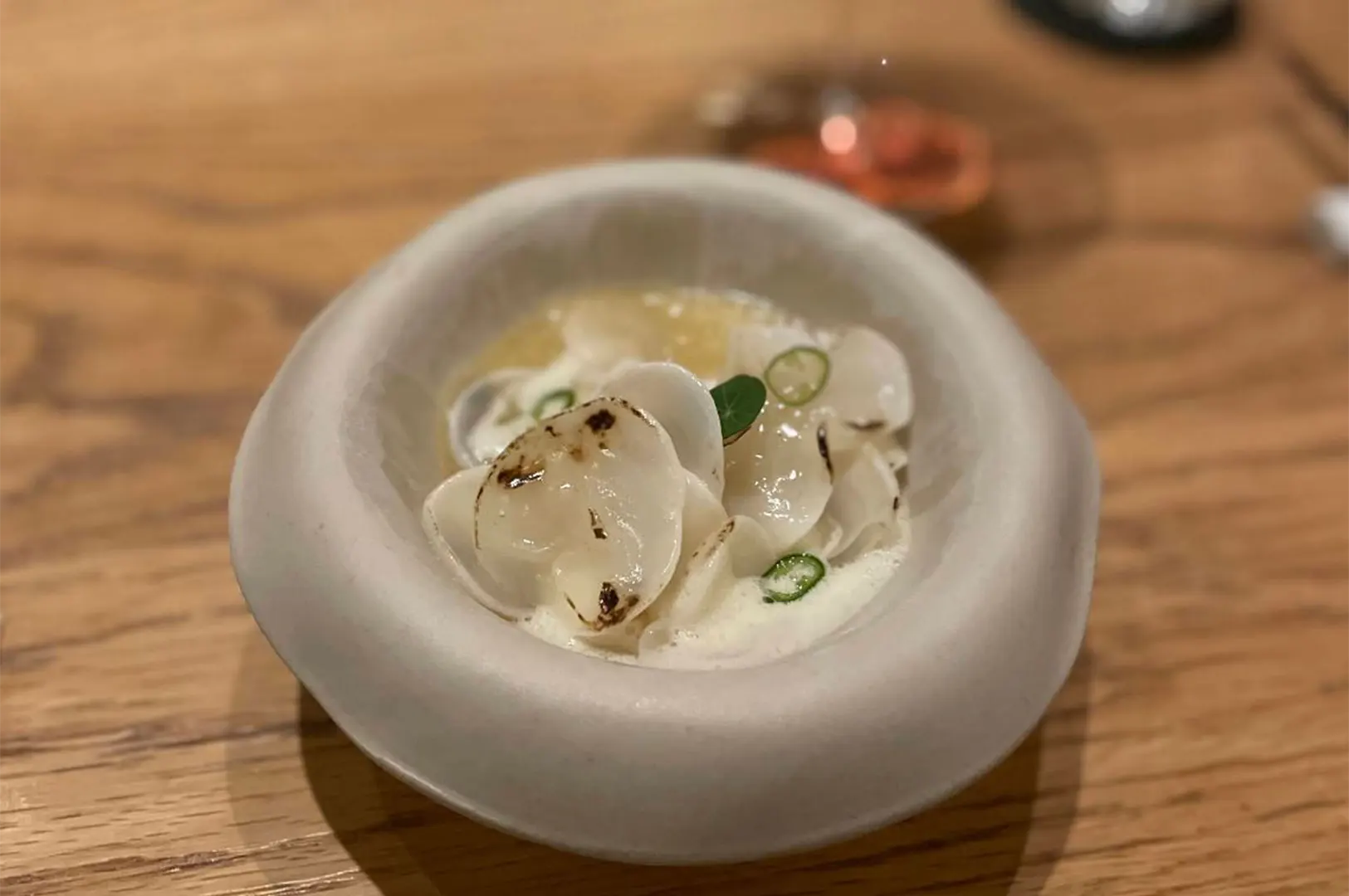 Delicately charred, paper-thin radish slices are artfully arranged over a light cream broth inside a smooth, beige ceramic bowl. Thinly sliced green chilies garnish the refined culinary dish, which rests on a natural wood table.