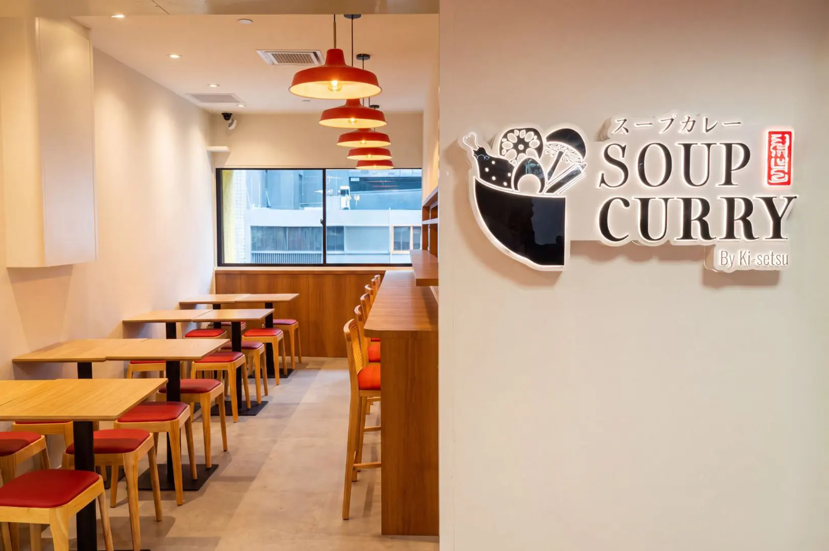 A modern restaurant entryway features a white wall with a raised "Soup Curry by Ki-setsu" logo alongside an illustrated bowl icon. Beyond the wall, a bright, minimalist dining area is lined with light wooden tables, red-cushioned seating, and hanging warm orange pendant lights.