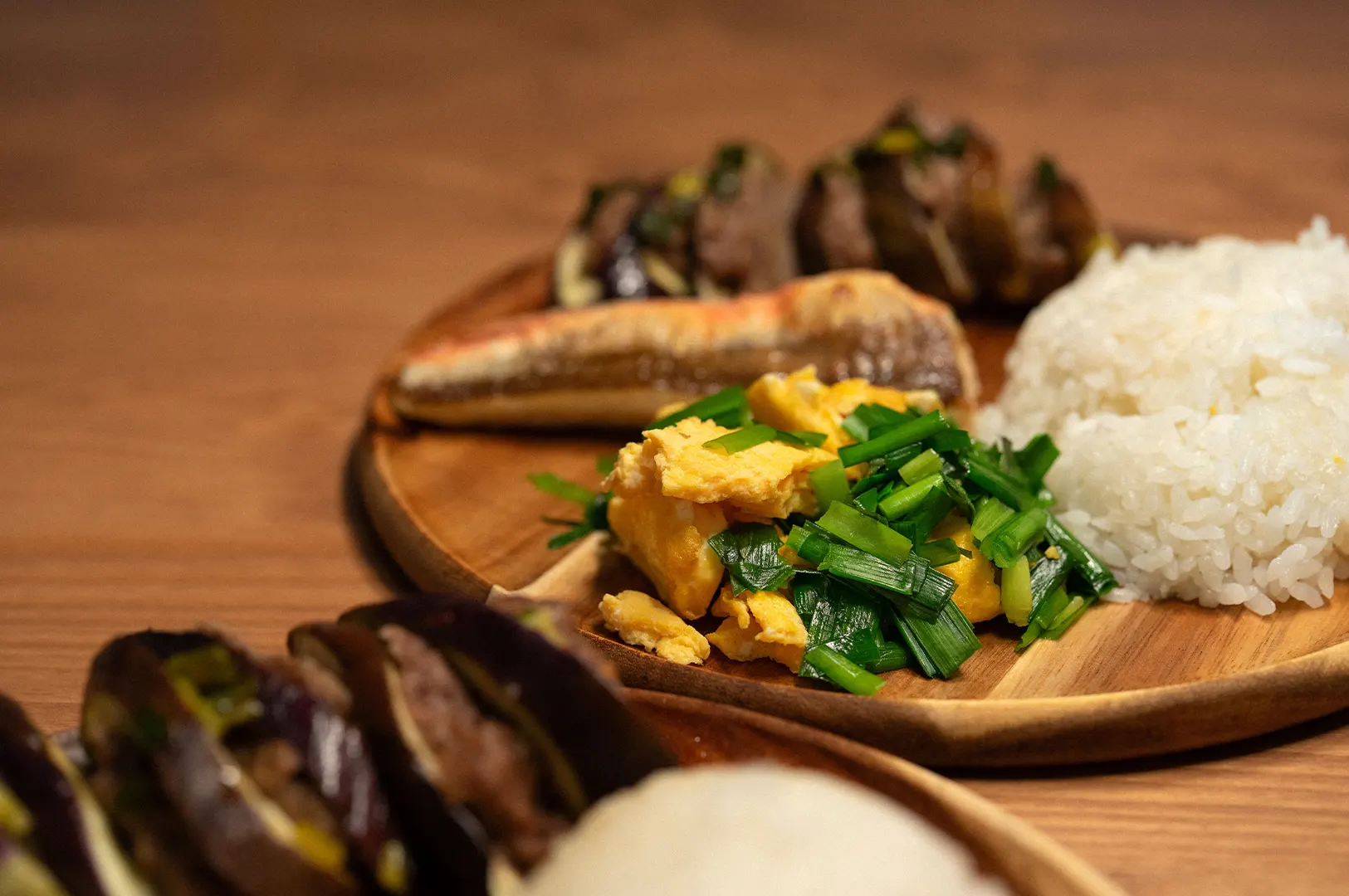 A round wooden platter holds a home-style meal featuring scrambled eggs garnished with chopped chives, steamed white rice, a grilled fish fillet, and stuffed eggplant slices. The warm-toned shot uses a shallow depth of field, bringing the scrambled eggs into sharp focus on a wooden table setting.