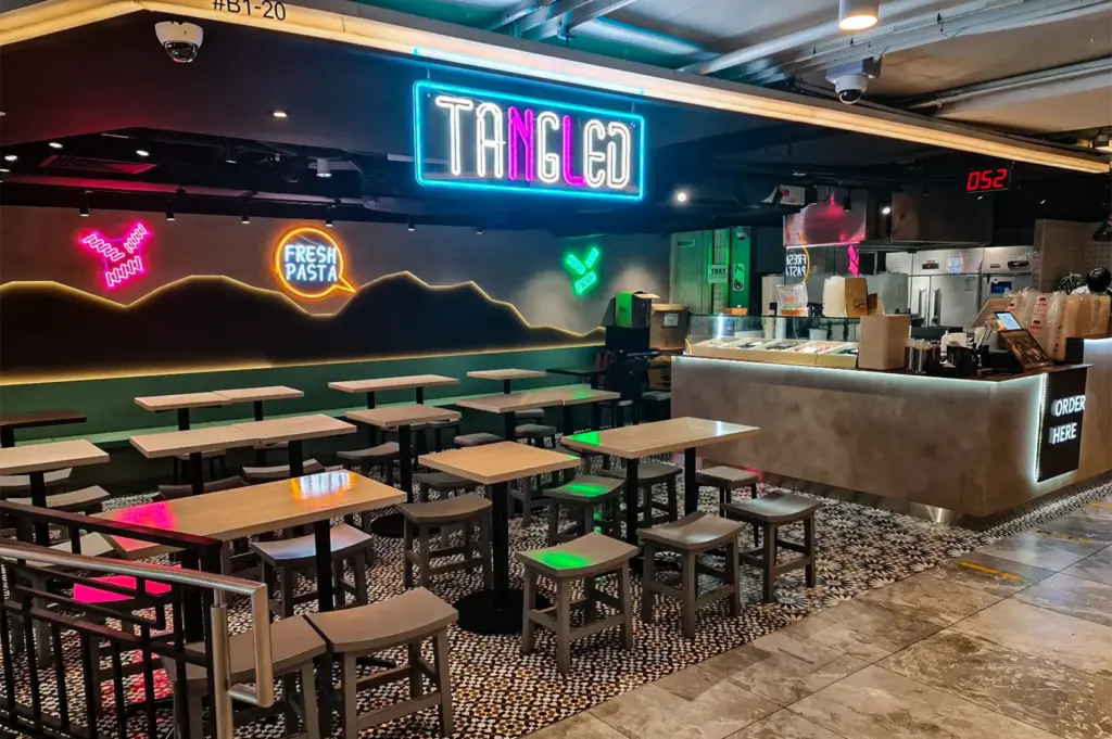 Bright cyan and pink neon signs reading "TANGLED" and "FRESH PASTA" illuminate the modern, empty dining area of a fast-casual eatery. Rows of light wood tables, small grey stools, and patterned tile flooring sit beside an illuminated order counter.