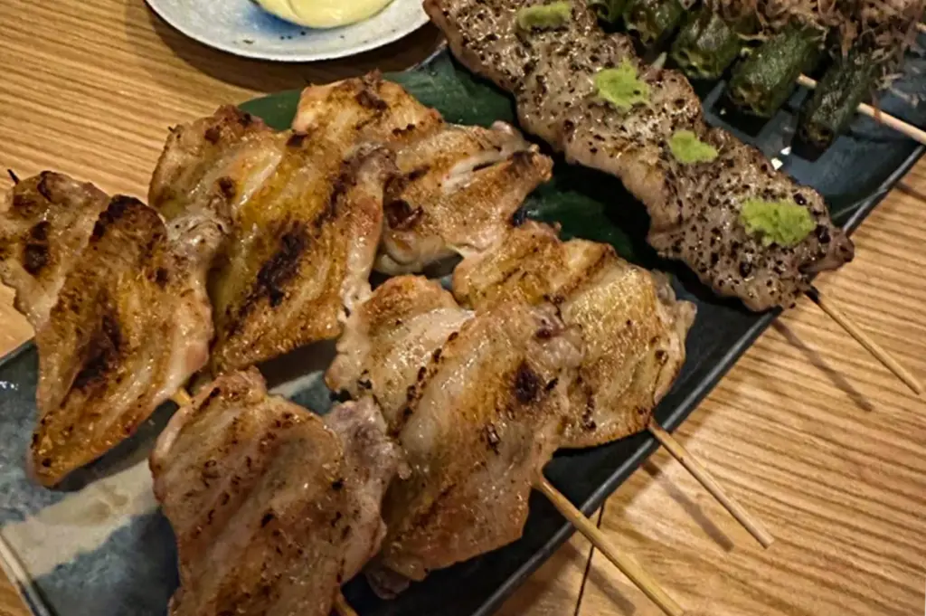 A high-angle, close-up shot captures an assortment of grilled yakitori skewers arranged on a long dark plate over a wooden table. The selection includes charred, golden-brown chicken wings on the left, alongside seasoned pork skewers topped with dollops of green wasabi paste and skewered vegetables in the background.