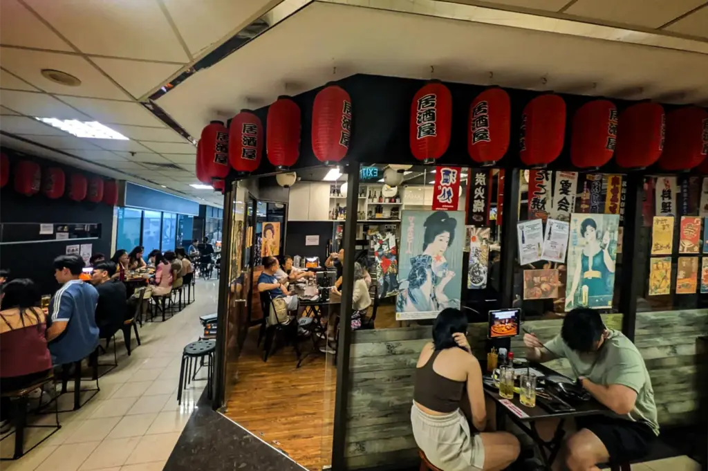 A high-angle shot captures patrons dining inside and outside a lively Japanese restaurant situated within a tiled indoor corridor. The eatery features a black facade wrapped in vibrant red paper lanterns, with the exterior and glass windows adorned with traditional banners and vintage Japanese illustrated posters.
