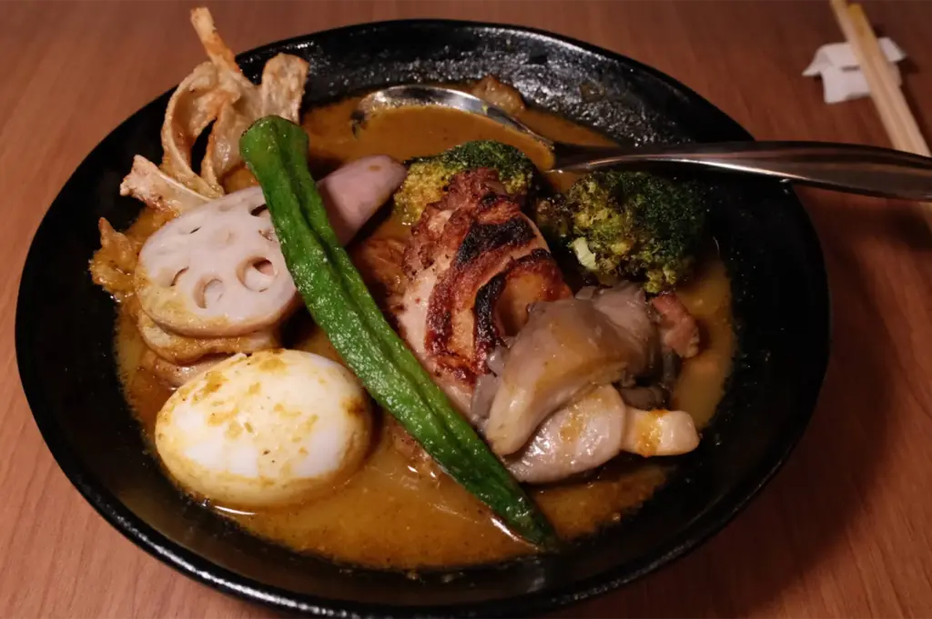 A close-up shot displays a dark ceramic bowl filled with rich Japanese soup curry, artfully topped with charred meat, a whole okra pod, sliced lotus root, broccoli, and a hard-boiled egg. A metal spoon rests inside the broth, with wooden chopsticks visible on the warm wooden tabletop in the background.