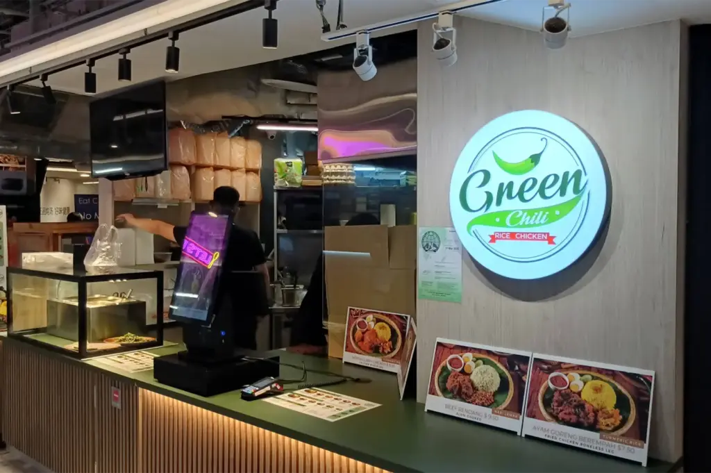 An illuminated circular sign reading "Green Chili Rice Chicken" hangs on a light wood wall above the order counter of a food stall. The counter features a self-ordering kiosk terminal, payment reader, and colorful food picture boards displaying dishes like Beef Rendang and Ayam Goreng Berempah.