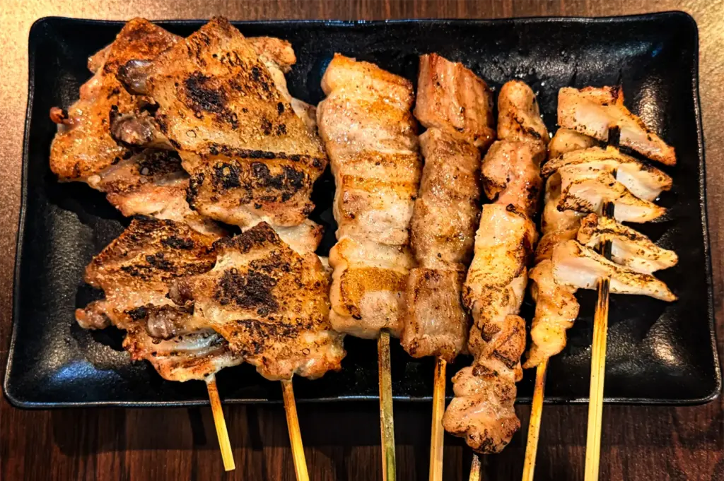 A close-up, top-down shot showcases an assortment of grilled Japanese yakitori skewers, including charred chicken wings and pork belly, arranged side-by-side. The glistening, seasoned skewers are presented together on a rectangular black plate over a dark wooden table.
