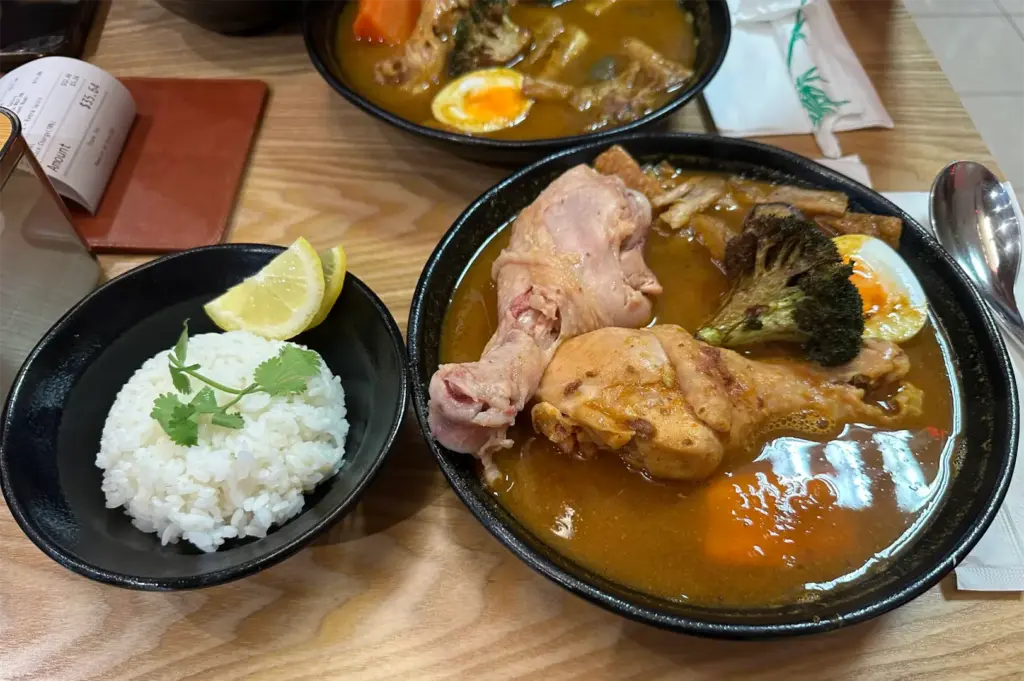 A broad bowl of Japanese soup curry features two whole chicken drumsticks, charred broccoli, and a soft-boiled egg immersed in savory broth. Alongside it rests a side bowl of molded white rice garnished with fresh cilantro and lemon wedges on a light wood table.