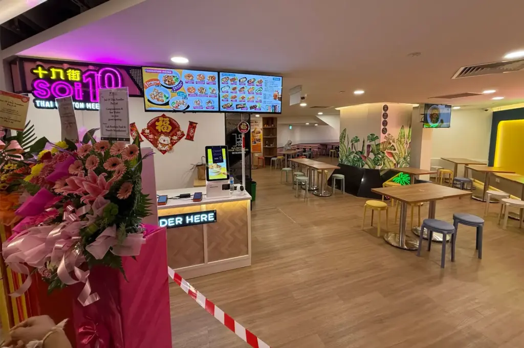 A bright pink floral arrangement stands near the entrance counter of a Thai restaurant called Soi 19, which features overhead digital menu displays and a self-ordering kiosk. The spacious dining area behind the counter is filled with wooden tables and colorful round stools under warm recessed lighting.