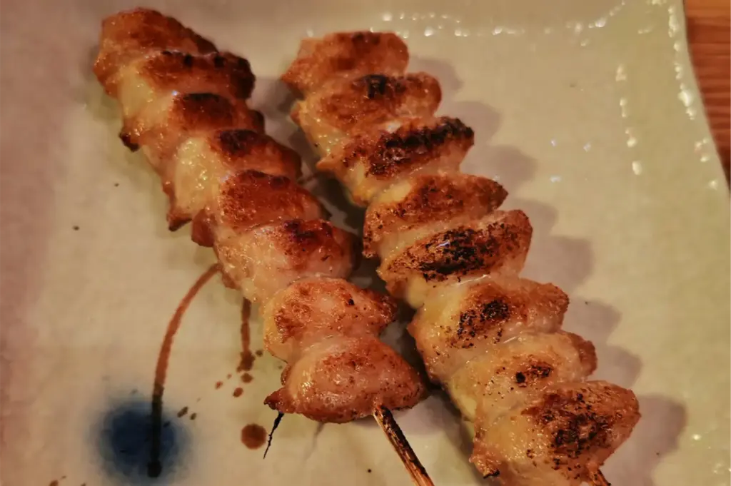 A close-up, high-angle shot captures two golden-brown, charred yakitori skewers resting diagonally on a textured ceramic plate. The grilled meat features crispy, glistening edges with distinct dark sear marks from the grill.