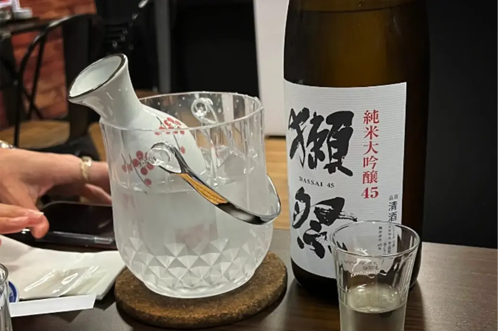 A close-up shot captures a bottle of Dassai 45 sake sitting next to an empty shot glass on a wooden table. To the left, a white ceramic sake carafe decorated with red floral patterns rests inside a textured, clear ice bucket filled with water and ice.